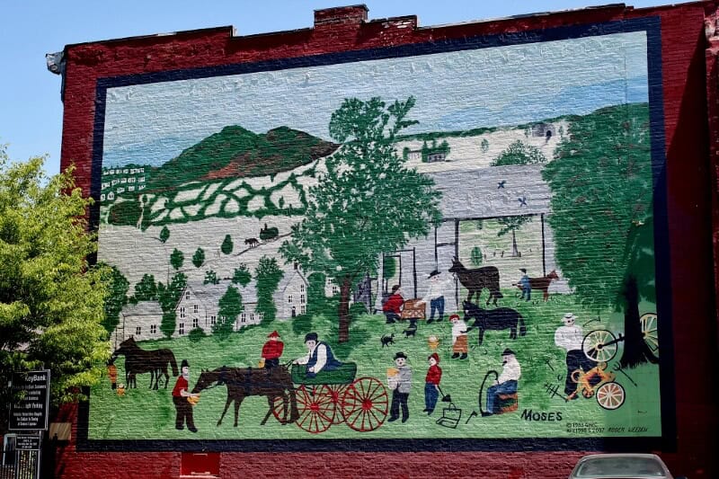 Mural of Grandma Moses painting showing  a building, people, and a horse and cart