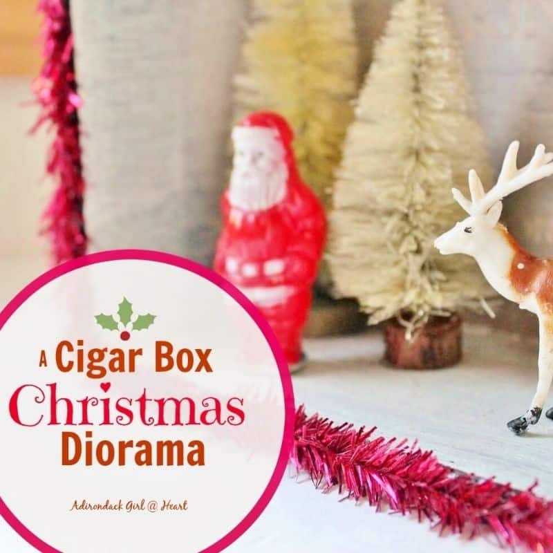 A cigar box Christmas diorama featuring a vintage Santa, white trees, and a deer